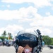 A toddler wearing an oversized military/aviation flight helmet with a dark visor stands outdoors at what appears to be an airshow or military open day event. The child is dressed in a pink floral dress, has a pink pacifier in her mouth, and is holding a white paper item. The helmet is far too large for the child, creating a humorous and endearing visual contrast.