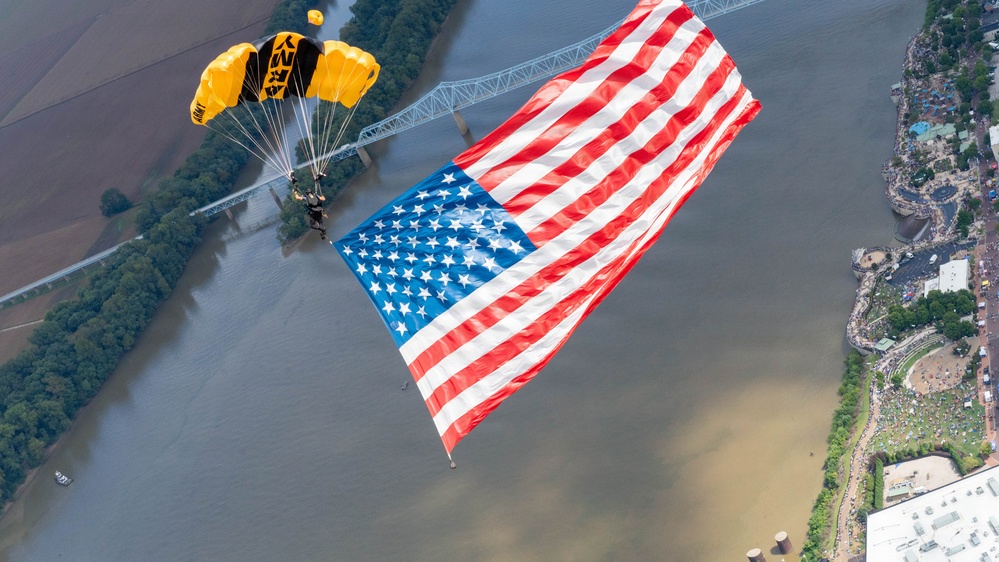 A military parachutist, identifiable by a yellow and black canopy marked 'ARMY', descends while carrying a very large American flag over a wide river. The aerial perspective reveals a bridge, tree-lined riverbanks, and what appears to be a large outdoor event or gathering on the right bank. The image captures a patriotic demonstration, likely part of an airshow or public ceremony.