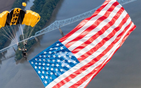 A military parachutist, identifiable by a yellow and black canopy marked 'ARMY', descends while carrying a very large American flag over a wide river. The aerial perspective reveals a bridge, tree-lined riverbanks, and what appears to be a large outdoor event or gathering on the right bank. The image captures a patriotic demonstration, likely part of an airshow or public ceremony.