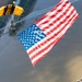 A military parachutist, identifiable by a yellow and black canopy marked 'ARMY', descends while carrying a very large American flag over a wide river. The aerial perspective reveals a bridge, tree-lined riverbanks, and what appears to be a large outdoor event or gathering on the right bank. The image captures a patriotic demonstration, likely part of an airshow or public ceremony.