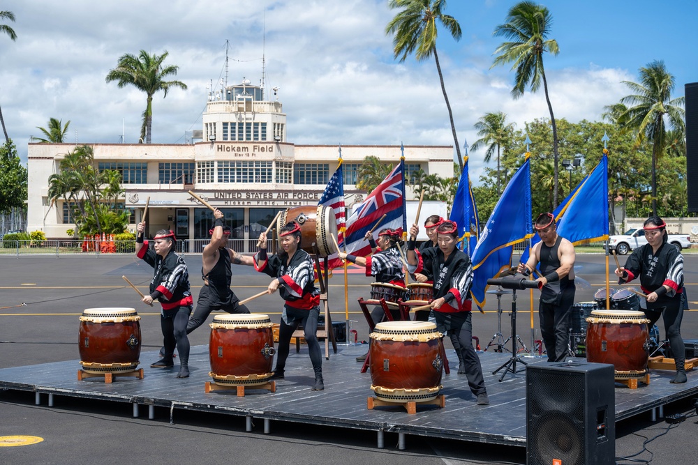 A taiko drumming ensemble performs on an outdoor stage at Hickam Field, Joint Base Pearl Harbor-Hickam, Hawaii. The performers, dressed in black uniforms with red accents and headbands, play traditional Japanese taiko drums in front of the historic United States Air Force Hickam Field building. Color guard members holding the American flag, Hawaii state flag, and U.S. Air Force flags stand behind the drummers.
