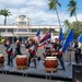 A taiko drumming ensemble performs on an outdoor stage at Hickam Field, Joint Base Pearl Harbor-Hickam, Hawaii. The performers, dressed in black uniforms with red accents and headbands, play traditional Japanese taiko drums in front of the historic United States Air Force Hickam Field building. Color guard members holding the American flag, Hawaii state flag, and U.S. Air Force flags stand behind the drummers.