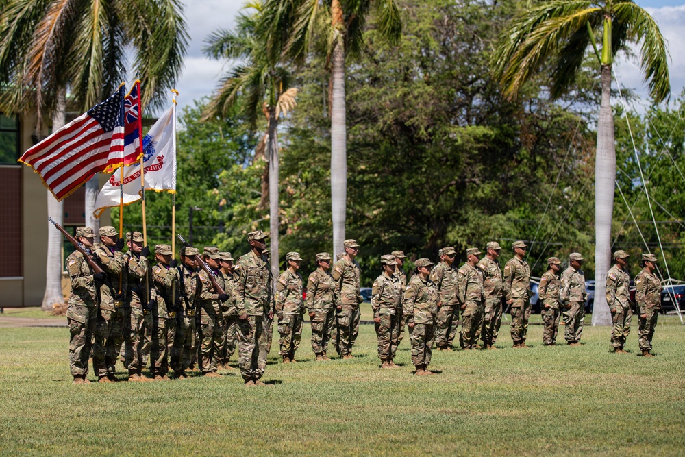 A military formation ceremony is taking place on a grass field in a tropical setting, likely Hawaii based on the palm trees and visible Hawaii state flag. Soldiers in Army OCP (Operational Camouflage Pattern) uniforms are standing in formation while a color guard presents multiple flags including the U.S. flag, Hawaii state flag, and at least one unit guidon. The scene appears to be a formal military ceremony such as a change of command, activation, or unit review.