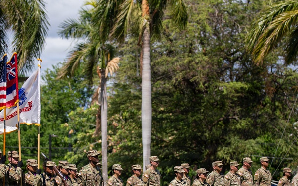 A military formation ceremony is taking place on a grass field in a tropical setting, likely Hawaii based on the palm trees and visible Hawaii state flag. Soldiers in Army OCP (Operational Camouflage Pattern) uniforms are standing in formation while a color guard presents multiple flags including the U.S. flag, Hawaii state flag, and at least one unit guidon. The scene appears to be a formal military ceremony such as a change of command, activation, or unit review.