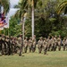 A military formation ceremony is taking place on a grass field in a tropical setting, likely Hawaii based on the palm trees and visible Hawaii state flag. Soldiers in Army OCP (Operational Camouflage Pattern) uniforms are standing in formation while a color guard presents multiple flags including the U.S. flag, Hawaii state flag, and at least one unit guidon. The scene appears to be a formal military ceremony such as a change of command, activation, or unit review.