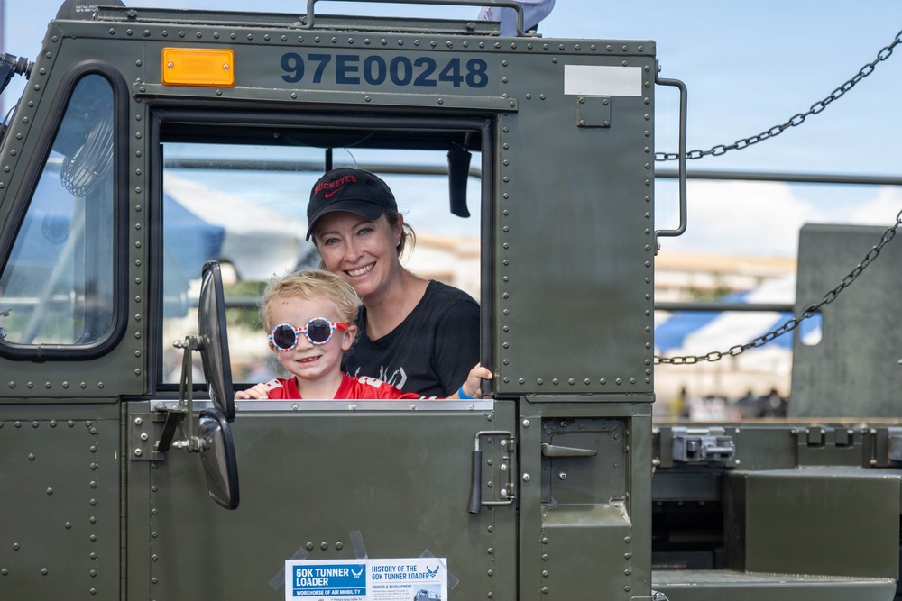 A woman and a young child are posing together inside the cab of a military olive-green vehicle identified as a 60K Tunner Loader (serial number 97E00248). The woman is wearing a black Nike 'Buckeyes' cap and black shirt, while the child wears a red shirt and patriotic star-patterned sunglasses. They appear to be at a public air show or military static display event.