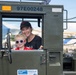 A woman and a young child are posing together inside the cab of a military olive-green vehicle identified as a 60K Tunner Loader (serial number 97E00248). The woman is wearing a black Nike 'Buckeyes' cap and black shirt, while the child wears a red shirt and patriotic star-patterned sunglasses. They appear to be at a public air show or military static display event.