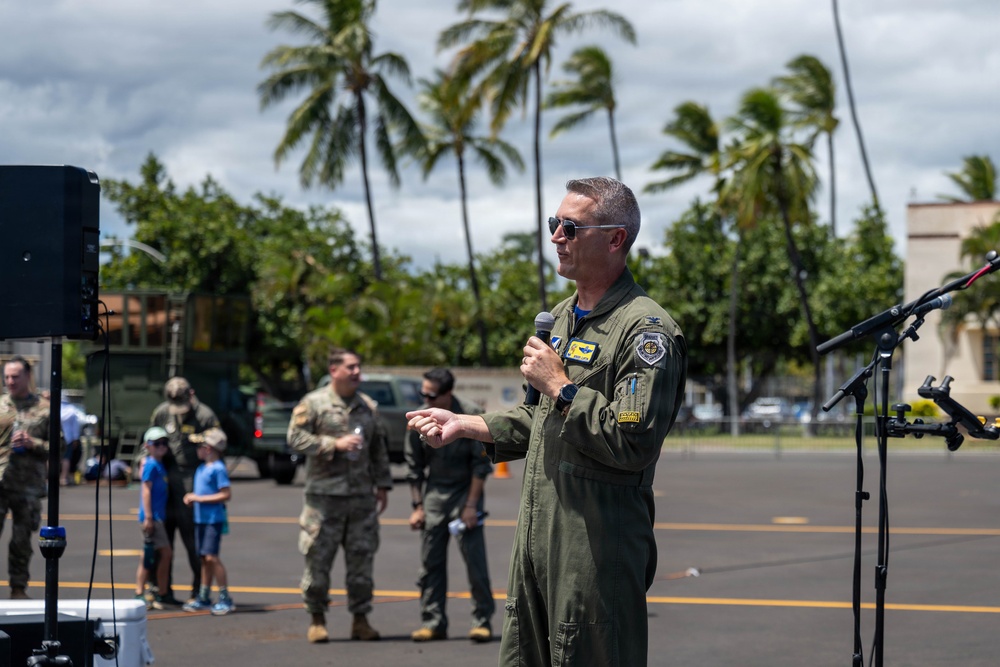 A military officer in an olive green flight suit is speaking into a microphone at an outdoor event, likely on a military base in a tropical location. He is wearing sunglasses and has visible patches on his uniform, suggesting a senior rank. Palm trees, cloudy skies, and other military personnel in the background indicate a formal outdoor ceremony or briefing.
