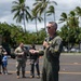 A military officer in an olive green flight suit is speaking into a microphone at an outdoor event, likely on a military base in a tropical location. He is wearing sunglasses and has visible patches on his uniform, suggesting a senior rank. Palm trees, cloudy skies, and other military personnel in the background indicate a formal outdoor ceremony or briefing.