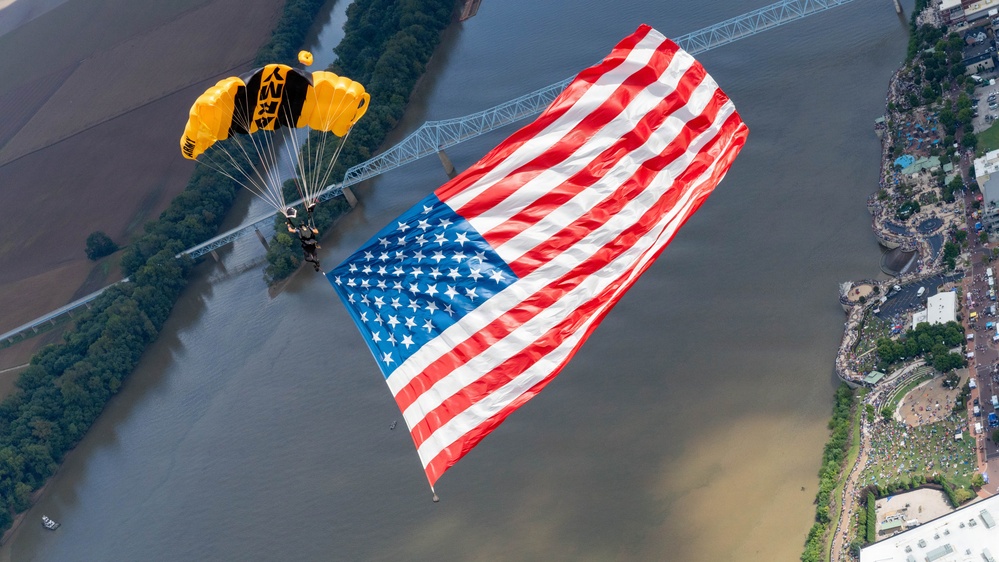 A military parachutist, identifiable by a yellow and black canopy with 'ARMY' markings, descends while carrying an enormous American flag over a river and urban landscape. The aerial perspective reveals a bridge, a large crowd gathered along the riverbank, and surrounding cityscape below. This appears to be a ceremonial demonstration jump, likely performed by the U.S. Army Golden Knights parachute team.