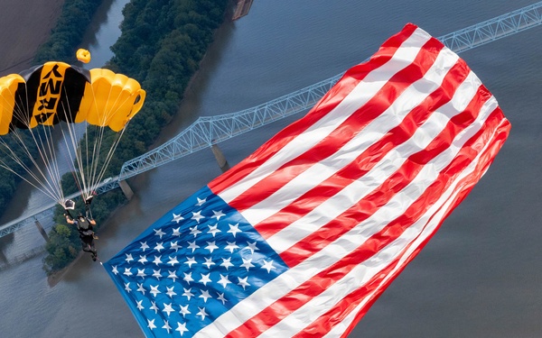 A military parachutist, identifiable by a yellow and black canopy with 'ARMY' markings, descends while carrying an enormous American flag over a river and urban landscape. The aerial perspective reveals a bridge, a large crowd gathered along the riverbank, and surrounding cityscape below. This appears to be a ceremonial demonstration jump, likely performed by the U.S. Army Golden Knights parachute team.