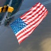 A military parachutist, identifiable by a yellow and black canopy with 'ARMY' markings, descends while carrying an enormous American flag over a river and urban landscape. The aerial perspective reveals a bridge, a large crowd gathered along the riverbank, and surrounding cityscape below. This appears to be a ceremonial demonstration jump, likely performed by the U.S. Army Golden Knights parachute team.