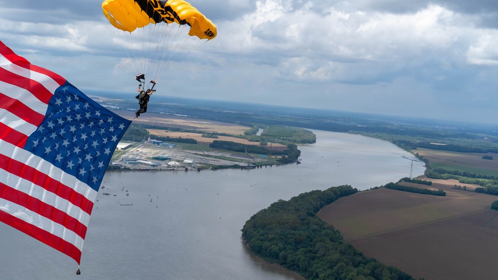 A military skydiver descends under a yellow and black parachute marked 'ARMY' while carrying a large American flag. The aerial photograph captures the jumper high above a wide river with agricultural fields, wooded peninsulas, and an industrial facility visible below. The overcast sky with scattered clouds provides a dramatic backdrop to this patriotic demonstration.