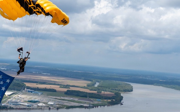 A military skydiver descends under a yellow and black parachute marked 'ARMY' while carrying a large American flag. The aerial photograph captures the jumper high above a wide river with agricultural fields, wooded peninsulas, and an industrial facility visible below. The overcast sky with scattered clouds provides a dramatic backdrop to this patriotic demonstration.