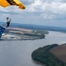A military skydiver descends under a yellow and black parachute marked 'ARMY' while carrying a large American flag. The aerial photograph captures the jumper high above a wide river with agricultural fields, wooded peninsulas, and an industrial facility visible below. The overcast sky with scattered clouds provides a dramatic backdrop to this patriotic demonstration.
