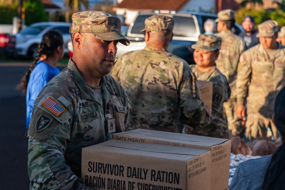 U.S. Army soldiers are distributing emergency relief supplies to civilians in what appears to be a disaster relief operation. The foreground soldier, identified by name tape as 'RAGRAGOLA,' is carrying a cardboard box labeled 'Survivor Daily Ration / Ración Diaria de Supervivencia.' Multiple soldiers in OCP (Operational Camouflage Pattern) uniforms are visible working together in an outdoor setting.