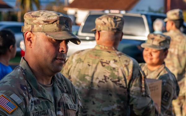 U.S. Army soldiers are distributing emergency relief supplies to civilians in what appears to be a disaster relief operation. The foreground soldier, identified by name tape as 'RAGRAGOLA,' is carrying a cardboard box labeled 'Survivor Daily Ration / Ración Diaria de Supervivencia.' Multiple soldiers in OCP (Operational Camouflage Pattern) uniforms are visible working together in an outdoor setting.