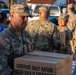 U.S. Army soldiers are distributing emergency relief supplies to civilians in what appears to be a disaster relief operation. The foreground soldier, identified by name tape as 'RAGRAGOLA,' is carrying a cardboard box labeled 'Survivor Daily Ration / Ración Diaria de Supervivencia.' Multiple soldiers in OCP (Operational Camouflage Pattern) uniforms are visible working together in an outdoor setting.