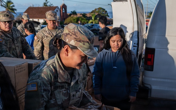 Military personnel and civilians are working together at what appears to be a disaster relief or food distribution operation. A soldier with the name tape 'MARTINEZ' in U.S. Army camouflage uniform is prominently handling cardboard boxes, while other uniformed soldiers and volunteers assist nearby. The setting appears to be an outdoor location near a white van, with multiple boxes of supplies being organized and distributed.