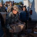 Military personnel and civilians are working together at what appears to be a disaster relief or food distribution operation. A soldier with the name tape 'MARTINEZ' in U.S. Army camouflage uniform is prominently handling cardboard boxes, while other uniformed soldiers and volunteers assist nearby. The setting appears to be an outdoor location near a white van, with multiple boxes of supplies being organized and distributed.