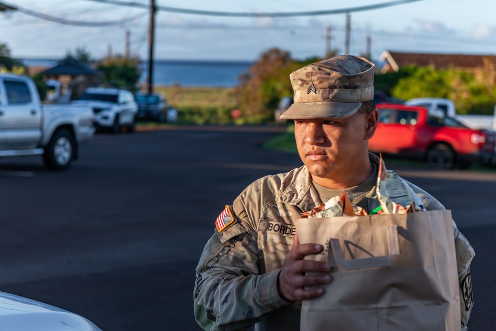 A U.S. Army Sergeant in MultiCam (OCP) uniform is holding a brown paper bag filled with items, likely participating in a supply or relief distribution effort. The soldier's name tape reads 'BORDE' and displays a Sergeant rank insignia on the patrol cap. The setting appears to be an outdoor parking area near a coastal location.
