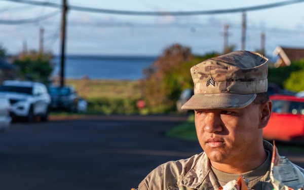 A U.S. Army Sergeant in MultiCam (OCP) uniform is holding a brown paper bag filled with items, likely participating in a supply or relief distribution effort. The soldier's name tape reads 'BORDE' and displays a Sergeant rank insignia on the patrol cap. The setting appears to be an outdoor parking area near a coastal location.