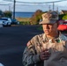 A U.S. Army Sergeant in MultiCam (OCP) uniform is holding a brown paper bag filled with items, likely participating in a supply or relief distribution effort. The soldier's name tape reads 'BORDE' and displays a Sergeant rank insignia on the patrol cap. The setting appears to be an outdoor parking area near a coastal location.