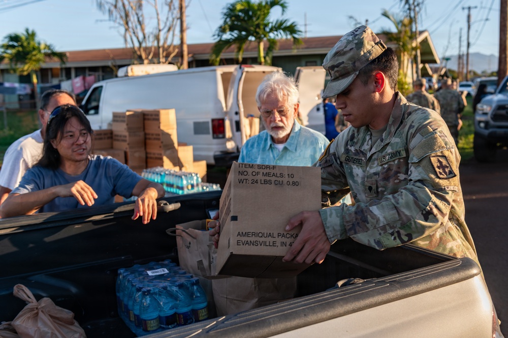 A U.S. Army soldier with the name tag 'Gutierrez' is distributing emergency meal rations (MRE-style packages) from the bed of a pickup truck to civilians. The box visible reads 'SDR-F, NO. OF MEALS: 10, WT: 24 LBS' and is labeled 'AMERIQUAL PACKA[GING], EVANSVILLE, IN.' The setting appears to be a disaster relief distribution point, likely in Hawaii based on the tropical vegetation and architecture.