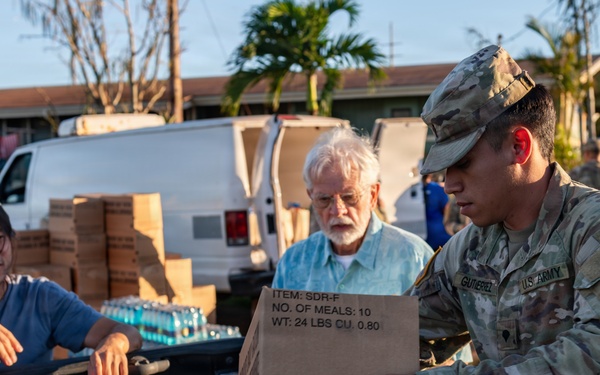 A U.S. Army soldier with the name tag 'Gutierrez' is distributing emergency meal rations (MRE-style packages) from the bed of a pickup truck to civilians. The box visible reads 'SDR-F, NO. OF MEALS: 10, WT: 24 LBS' and is labeled 'AMERIQUAL PACKA[GING], EVANSVILLE, IN.' The setting appears to be a disaster relief distribution point, likely in Hawaii based on the tropical vegetation and architecture.
