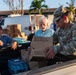 A U.S. Army soldier with the name tag 'Gutierrez' is distributing emergency meal rations (MRE-style packages) from the bed of a pickup truck to civilians. The box visible reads 'SDR-F, NO. OF MEALS: 10, WT: 24 LBS' and is labeled 'AMERIQUAL PACKA[GING], EVANSVILLE, IN.' The setting appears to be a disaster relief distribution point, likely in Hawaii based on the tropical vegetation and architecture.