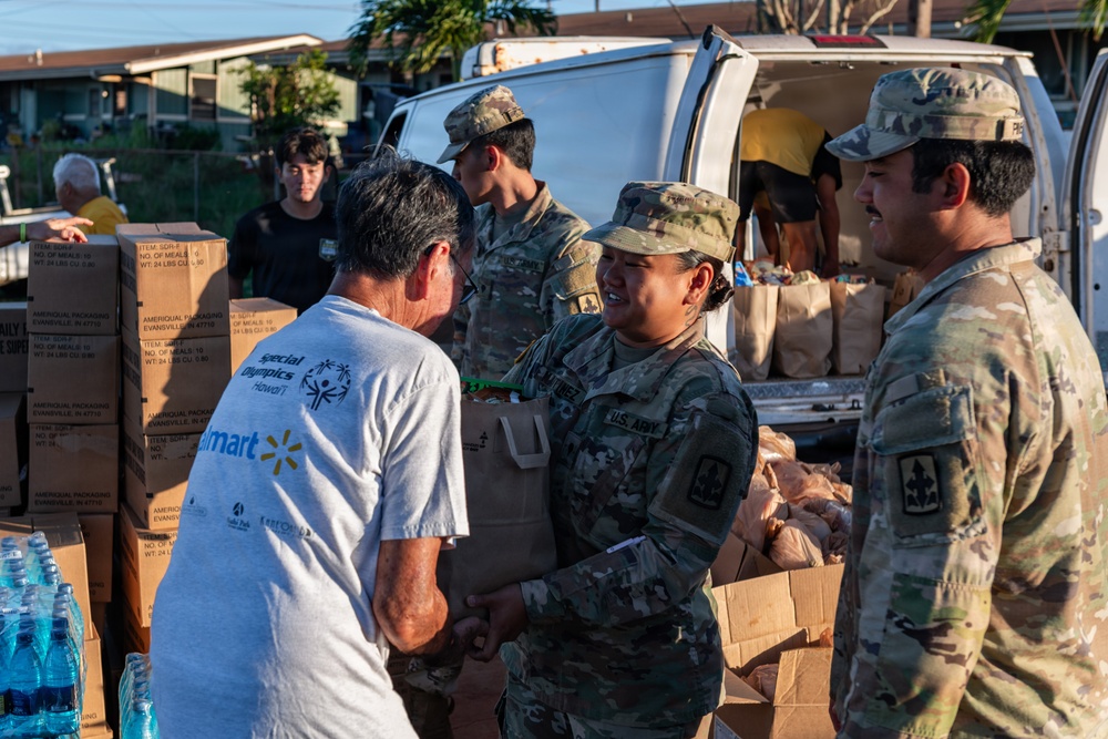 U.S. Army soldiers in MultiCam camouflage uniforms are distributing food and supplies to civilians at what appears to be a disaster relief or humanitarian aid distribution point. A female soldier is handing a bag of goods to a civilian wearing a Special Olympics Hawaii and Walmart t-shirt. Multiple cardboard boxes, water bottles, and paper bags filled with supplies are visible around a white van.