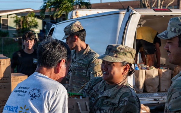 U.S. Army soldiers in MultiCam camouflage uniforms are distributing food and supplies to civilians at what appears to be a disaster relief or humanitarian aid distribution point. A female soldier is handing a bag of goods to a civilian wearing a Special Olympics Hawaii and Walmart t-shirt. Multiple cardboard boxes, water bottles, and paper bags filled with supplies are visible around a white van.