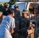 U.S. Army soldiers in MultiCam camouflage uniforms are distributing food and supplies to civilians at what appears to be a disaster relief or humanitarian aid distribution point. A female soldier is handing a bag of goods to a civilian wearing a Special Olympics Hawaii and Walmart t-shirt. Multiple cardboard boxes, water bottles, and paper bags filled with supplies are visible around a white van.