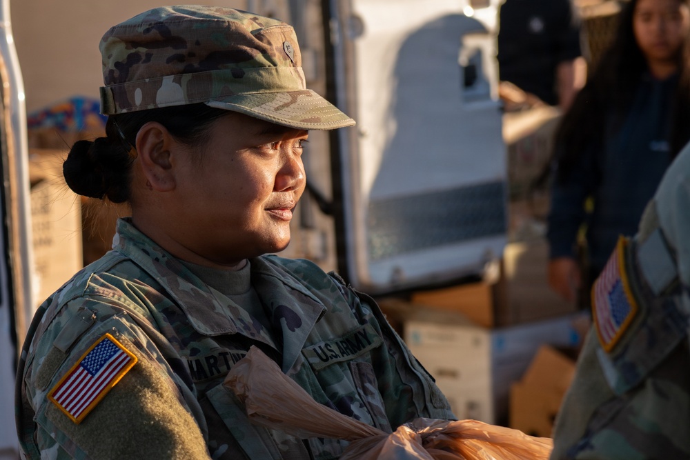 A U.S. Army soldier, identified by the name tape 'MARTINEZ' and wearing Operational Camouflage Pattern (OCP) uniform, is engaged in what appears to be a food or supply distribution activity. The soldier is wearing an American flag patch on the left shoulder and a U.S. Army tape on the chest. The setting appears to be an outdoor distribution event with boxes and supplies visible in the background.