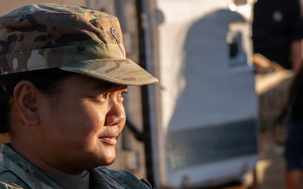 A U.S. Army soldier, identified by the name tape 'MARTINEZ' and wearing Operational Camouflage Pattern (OCP) uniform, is engaged in what appears to be a food or supply distribution activity. The soldier is wearing an American flag patch on the left shoulder and a U.S. Army tape on the chest. The setting appears to be an outdoor distribution event with boxes and supplies visible in the background.