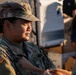 A U.S. Army soldier, identified by the name tape 'MARTINEZ' and wearing Operational Camouflage Pattern (OCP) uniform, is engaged in what appears to be a food or supply distribution activity. The soldier is wearing an American flag patch on the left shoulder and a U.S. Army tape on the chest. The setting appears to be an outdoor distribution event with boxes and supplies visible in the background.
