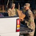 Two U.S. Army soldiers in MultiCam/OCP camouflage uniforms are unloading or distributing supplies from the bed of a white pickup truck. One soldier is carrying a cardboard box labeled 'NO. OF MEALS: 10' and 'WT: 24 LBS', indicating disaster relief or humanitarian aid operations. The setting appears to be a residential area with visible storm or disaster damage in the background.