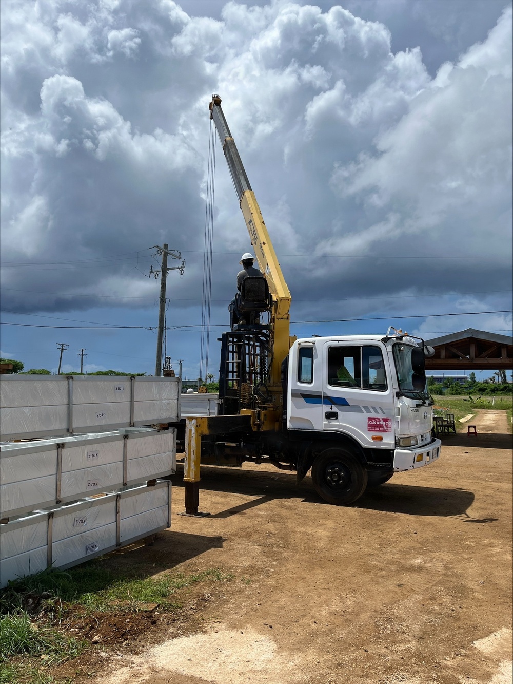 A white Hyundai truck-mounted crane with a yellow boom arm is actively operating at a construction or delivery site. A worker wearing a white hard hat is seated at the crane controls, apparently lifting or positioning stacked precast concrete or insulated wall panels. The scene is set on a dirt lot under a dramatic partly cloudy sky.