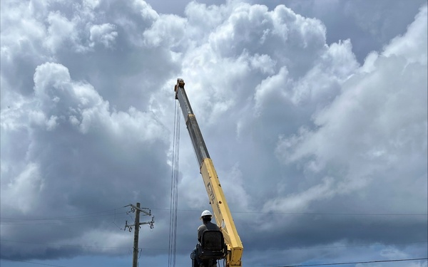A white Hyundai truck-mounted crane with a yellow boom arm is actively operating at a construction or delivery site. A worker wearing a white hard hat is seated at the crane controls, apparently lifting or positioning stacked precast concrete or insulated wall panels. The scene is set on a dirt lot under a dramatic partly cloudy sky.