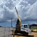 A white Hyundai truck-mounted crane with a yellow boom arm is actively operating at a construction or delivery site. A worker wearing a white hard hat is seated at the crane controls, apparently lifting or positioning stacked precast concrete or insulated wall panels. The scene is set on a dirt lot under a dramatic partly cloudy sky.