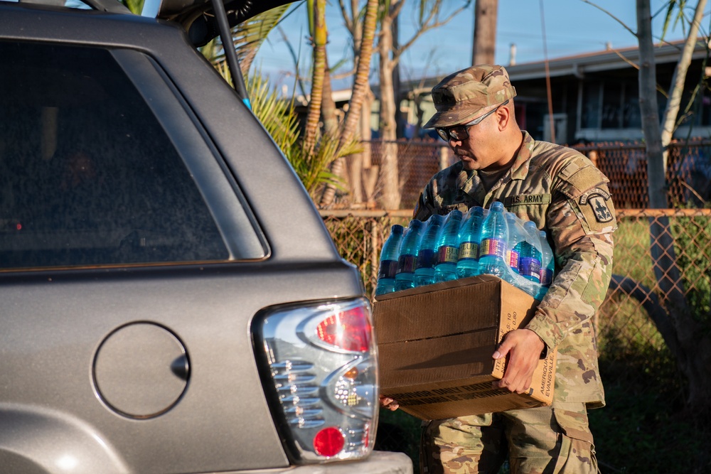 A U.S. Army soldier in camouflage uniform and cap is loading or unloading a case of bottled water into the trunk of a gray SUV. The setting appears to be a residential neighborhood, suggesting a disaster relief or humanitarian aid distribution operation. The cardboard box contains multiple plastic water bottles and bears partial text referencing 'America' and 'Evansville'.