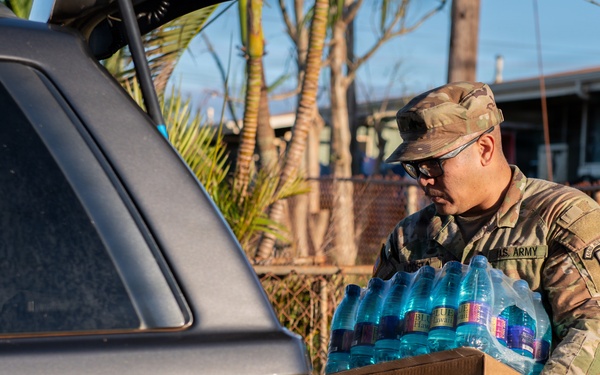 A U.S. Army soldier in camouflage uniform and cap is loading or unloading a case of bottled water into the trunk of a gray SUV. The setting appears to be a residential neighborhood, suggesting a disaster relief or humanitarian aid distribution operation. The cardboard box contains multiple plastic water bottles and bears partial text referencing 'America' and 'Evansville'.