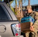 A U.S. Army soldier in camouflage uniform and cap is loading or unloading a case of bottled water into the trunk of a gray SUV. The setting appears to be a residential neighborhood, suggesting a disaster relief or humanitarian aid distribution operation. The cardboard box contains multiple plastic water bottles and bears partial text referencing 'America' and 'Evansville'.