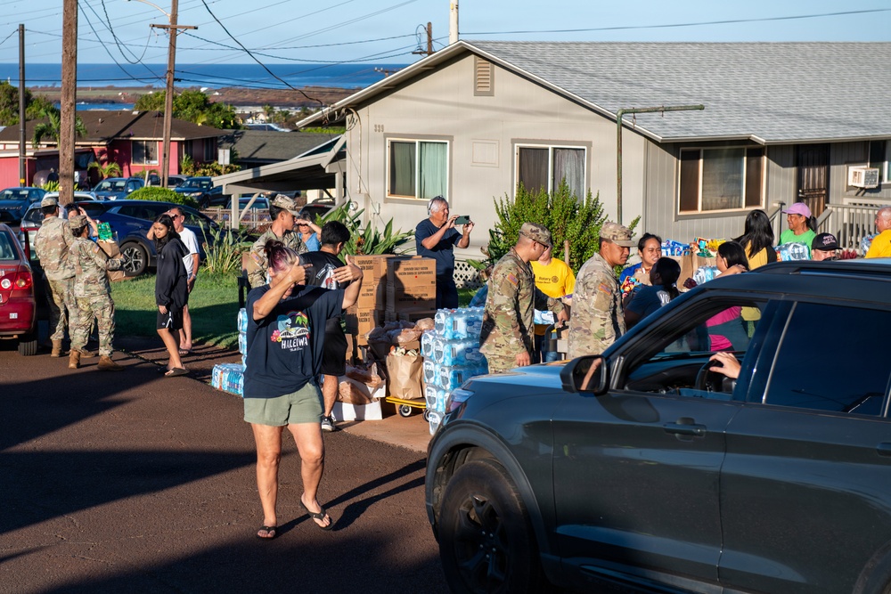 A community relief distribution event is taking place on a residential street, with military personnel in camouflage uniforms working alongside civilians to distribute supplies. Large quantities of bottled water cases and cardboard boxes are staged in the area, being handed out to people in vehicles and on foot. The setting appears to be a Hawaiian neighborhood based on visible signage and coastal backdrop.