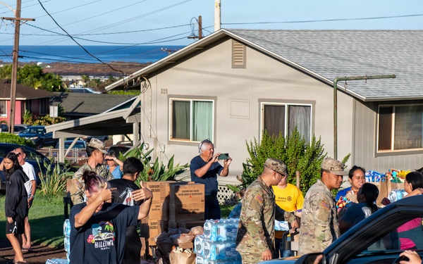 A community relief distribution event is taking place on a residential street, with military personnel in camouflage uniforms working alongside civilians to distribute supplies. Large quantities of bottled water cases and cardboard boxes are staged in the area, being handed out to people in vehicles and on foot. The setting appears to be a Hawaiian neighborhood based on visible signage and coastal backdrop.