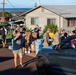 A community relief distribution event is taking place on a residential street, with military personnel in camouflage uniforms working alongside civilians to distribute supplies. Large quantities of bottled water cases and cardboard boxes are staged in the area, being handed out to people in vehicles and on foot. The setting appears to be a Hawaiian neighborhood based on visible signage and coastal backdrop.