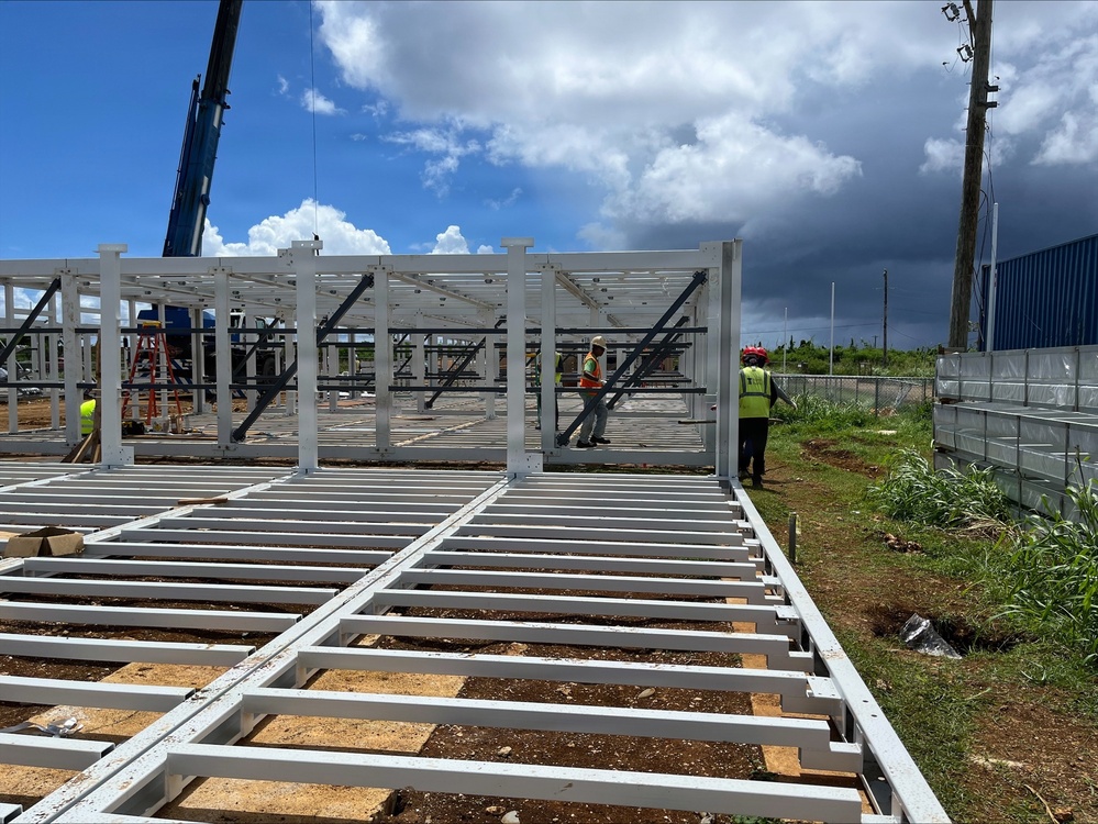 A construction site showing the early structural assembly of a steel-framed building. Workers in high-visibility vests are actively assembling white-painted steel columns and floor joists on a cleared dirt lot. A crane is visible in the background, and an existing blue metal building stands nearby.