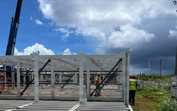 A construction site showing the early structural assembly of a steel-framed building. Workers in high-visibility vests are actively assembling white-painted steel columns and floor joists on a cleared dirt lot. A crane is visible in the background, and an existing blue metal building stands nearby.