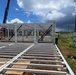 A construction site showing the early structural assembly of a steel-framed building. Workers in high-visibility vests are actively assembling white-painted steel columns and floor joists on a cleared dirt lot. A crane is visible in the background, and an existing blue metal building stands nearby.