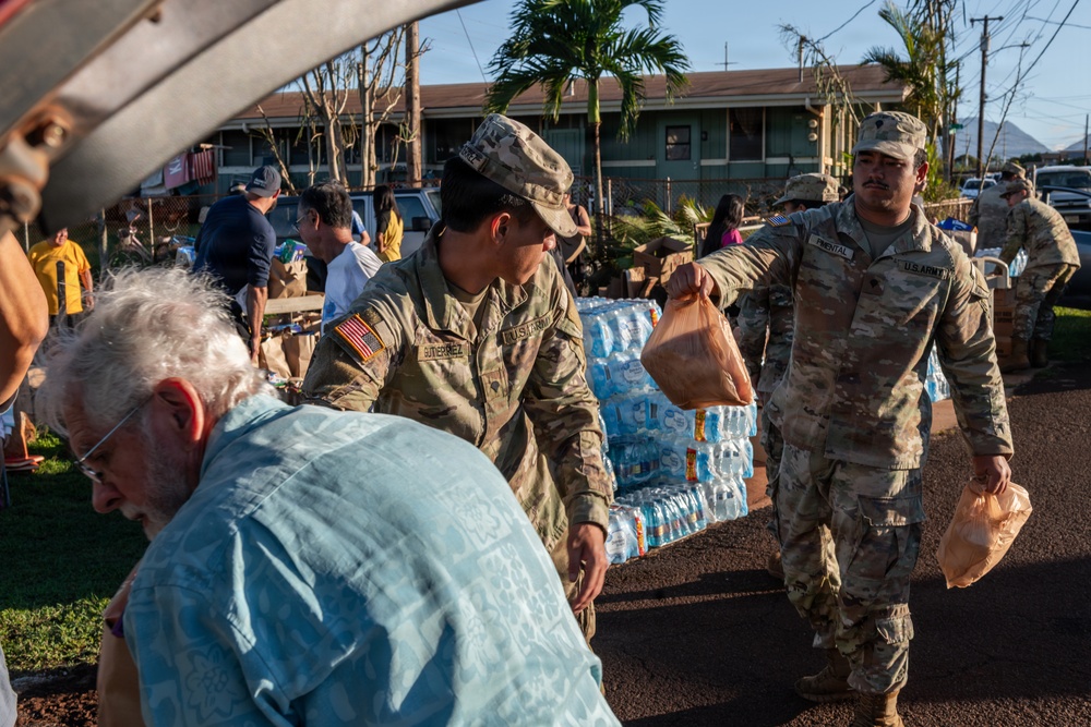 U.S. Army soldiers are distributing relief supplies, including bottled water and food bags, to civilians at what appears to be a disaster relief distribution point. Multiple soldiers in OCP (Operational Camouflage Pattern) uniforms with U.S. Army patches are actively handing out supplies. Large pallets of bottled water are visible in the background, suggesting a organized emergency relief operation.